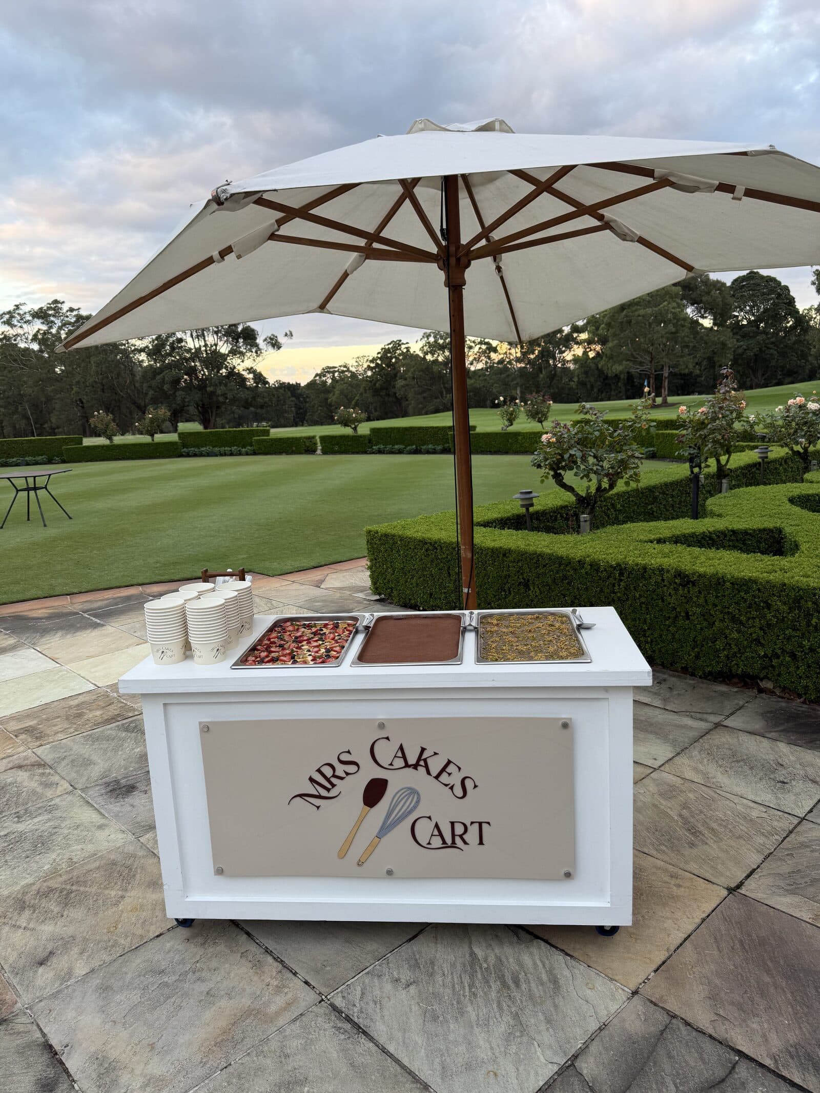 A Mrs Cakes cart set up with three trays of dessert at a garden venue