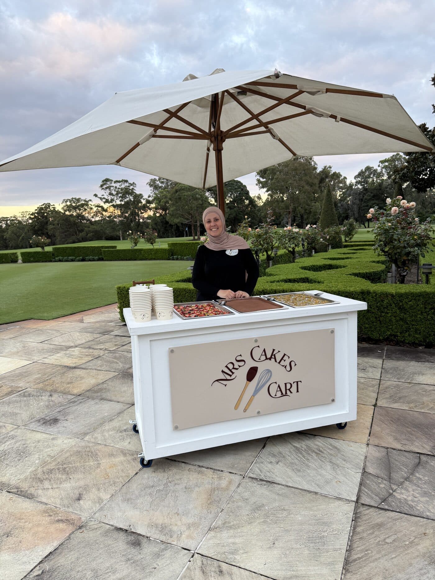 Sara standing behind her cake cart under a white umbrella at a garden venue